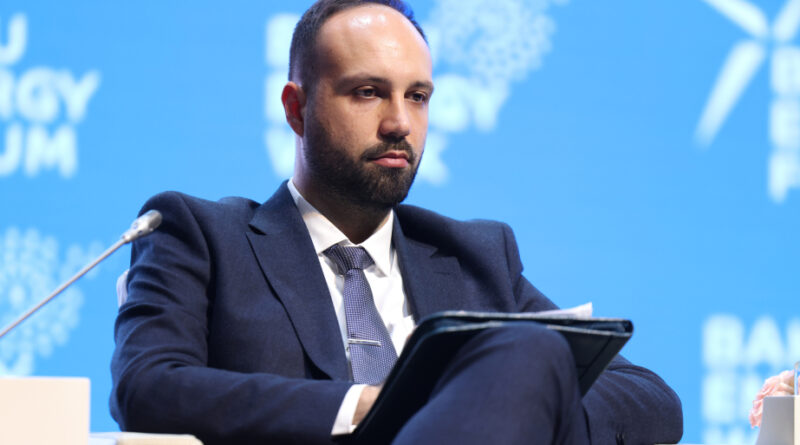 Male panelist in a dark blue suit and tie seated on stage with a tablet against a blue conference backdrop.