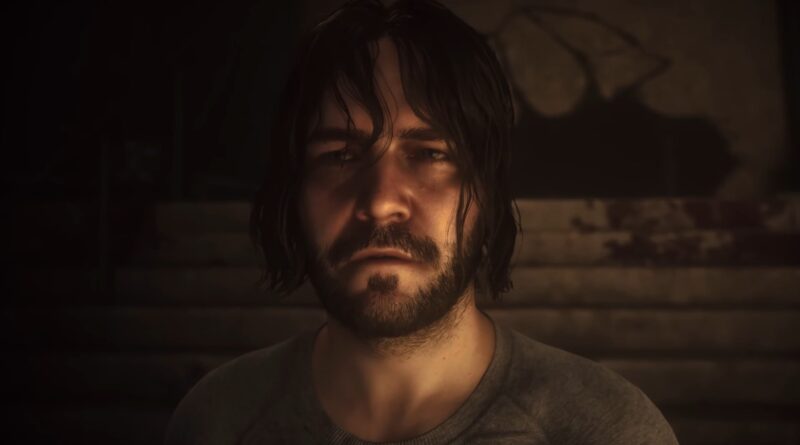 Close-up portrait of a man with dark hair and beard in a dim, moody light, looking toward the camera.