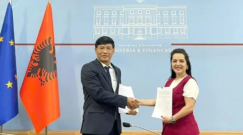 Two professionals shake hands after signing documents in a formal setting, with EU and Albania flags nearby and a blue backdrop showing a building outline.