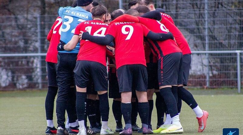 A group of FC Albania players in red jerseys form a tight huddle on a green soccer field, arms around each other before a game.