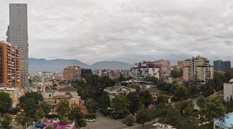 City skyline with a mix of tall and mid-rise buildings, trees, and distant mountains under a cloudy sky.