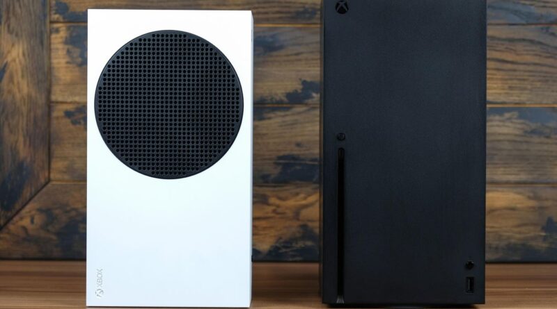 Two Xbox consoles sit side by side on a wooden surface: a white model with a large circular vent on the front left and a tall black model on the right showng a sleek, solid back panel.
