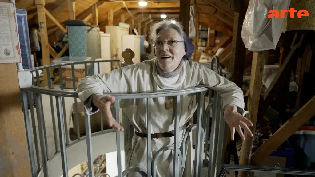 Smiling elderly woman leaning through a metal railing in a cluttered attic storage space, reaching toward the camera.
