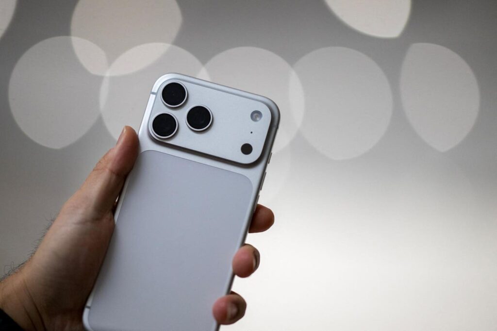 Person holding a silver iPhone with a triple-camera module on the back, shown at an angle with a soft bokeh background