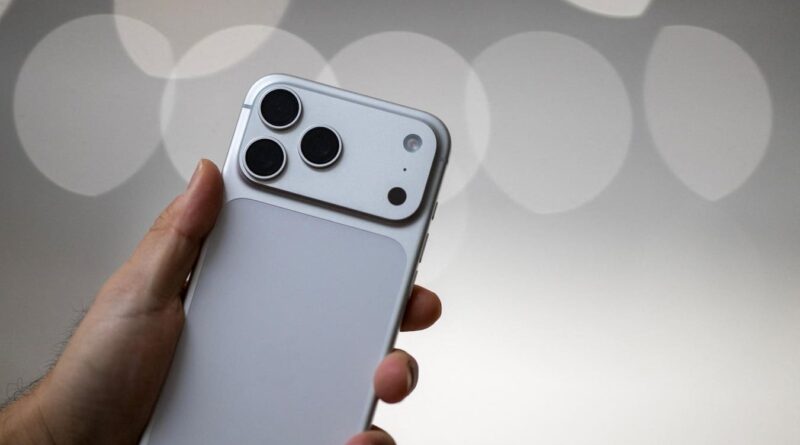 Person holding a silver iPhone with a triple-camera module on the back, shown at an angle with a soft bokeh background