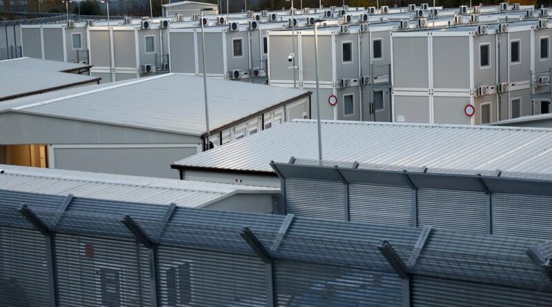 Row of gray modular buildings with air conditioning units on the roofs, viewed from behind a tall metal fence in a secured compound.