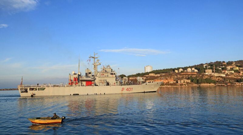 Military ship P401 anchored near a rocky coastline with buildings on a hill and a small orange boat in the foreground.