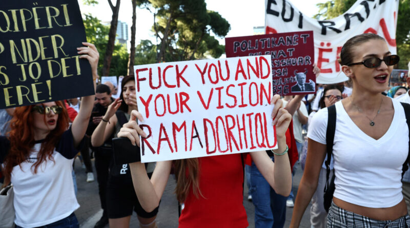 Protesters marching on a sunny street; center sign reads 'FUCK YOU AND YOUR VISION' amid other signs and banners.