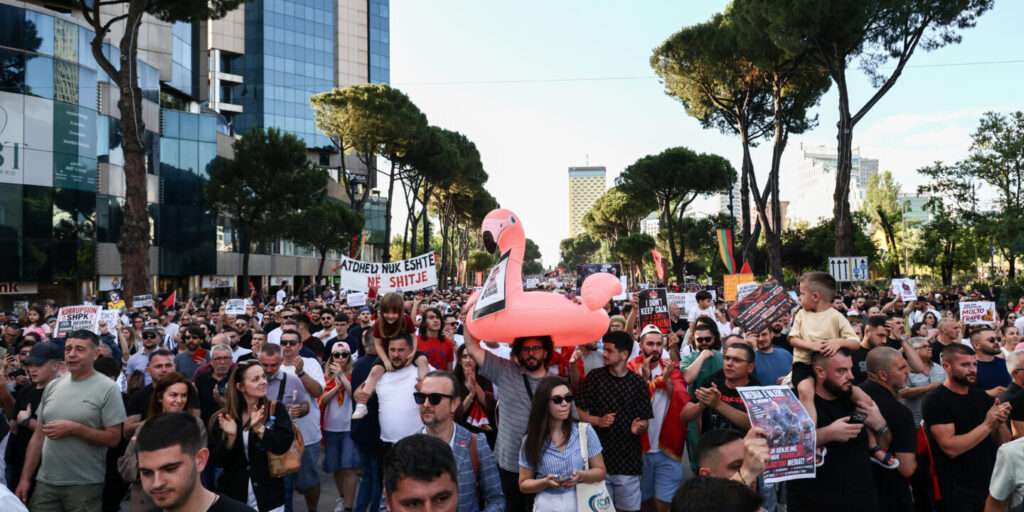 Large crowd marching in a city street with protest signs; a pink flamingo float is held above the heads of protesters.