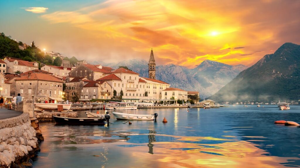 Sunset over a coastal town with red-roofed stone buildings, boats in a calm harbor, and distant mountains.