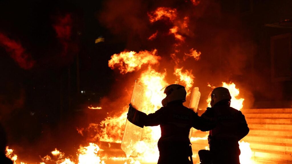 Two firefighters in helmets and protective gear stand before a huge orange blaze at night, holding shields up to defend against the flames.