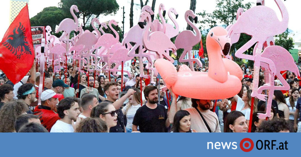Crowd of people at a street parade carrying oversized pink flamingo balloons; a red flag with a black eagle is visible on the left, and a blue banner with 'news ORF.at' runs across the bottom.