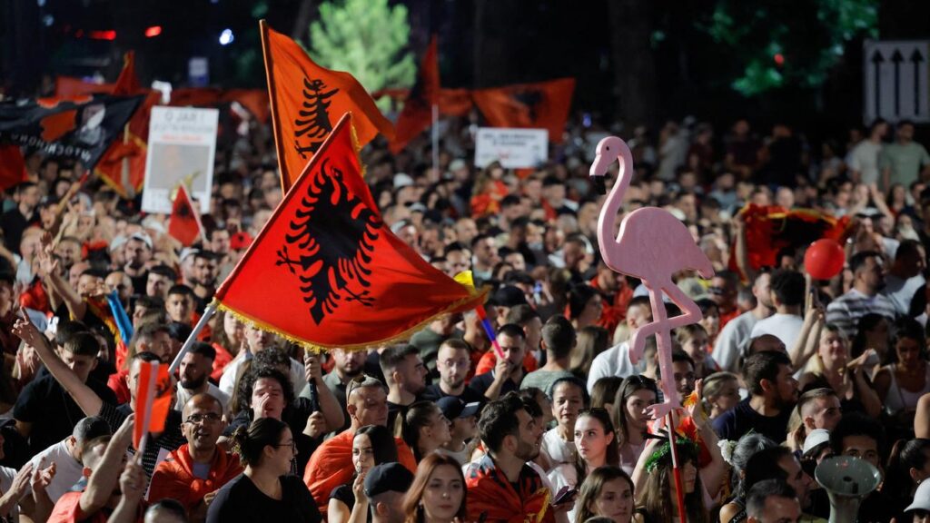 Dense nighttime crowd at a rally, many holding red Albanian flags with a black double-headed eagle and a pink flamingo cutout in the foreground.