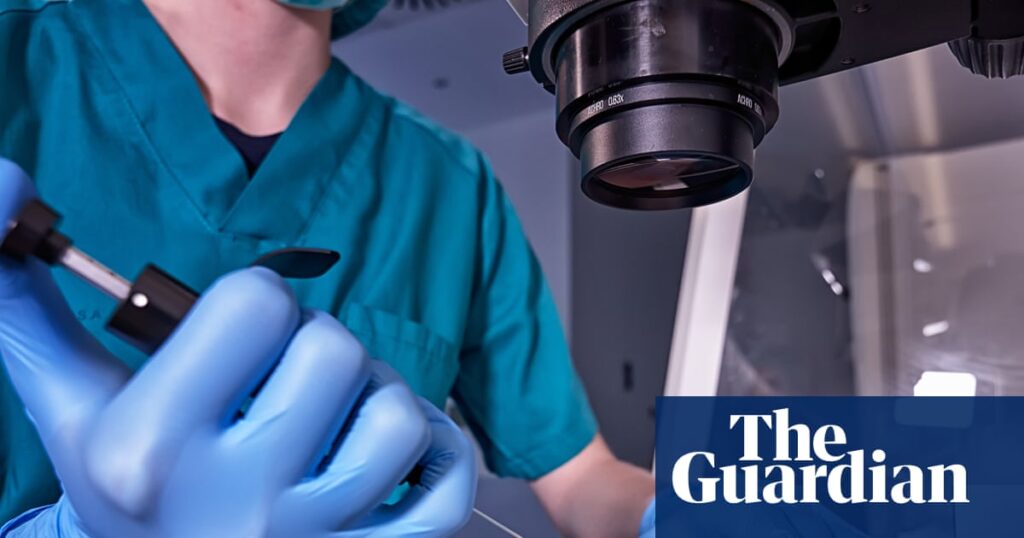 Medical researcher in teal scrubs and blue gloves operating a precision instrument beside a microscope.