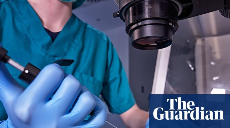 Medical researcher in teal scrubs and blue gloves operating a precision instrument beside a microscope.