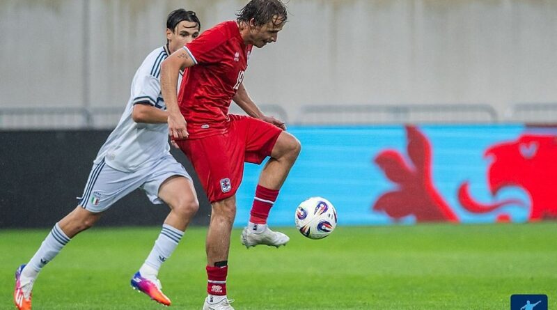Soccer players contesting for the ball on a rainy day; a red-kit player controls the ball while a white-kit defender closes in behind him on the green field.
