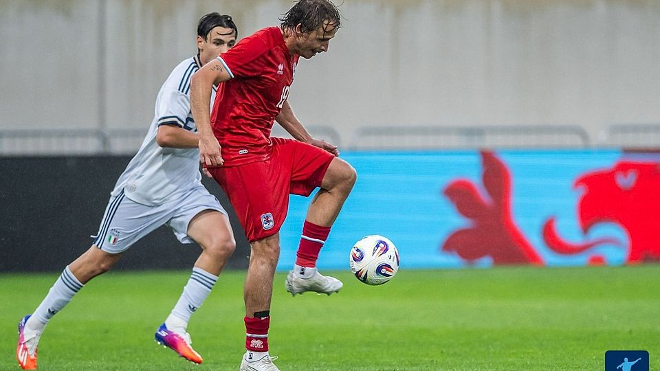 Soccer players contesting for the ball on a rainy day; a red-kit player controls the ball while a white-kit defender closes in behind him on the green field.