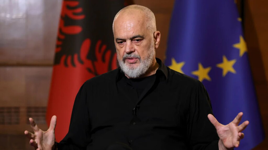 Older man with a gray beard speaks in an interview, wearing a black shirt, gesturing with his hands; EU flag visible in the background.