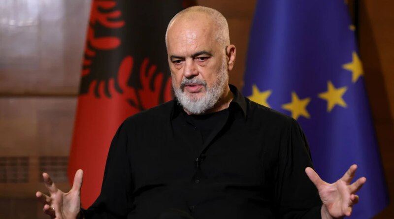 Older man with a gray beard speaks in an interview, wearing a black shirt, gesturing with his hands; EU flag visible in the background.