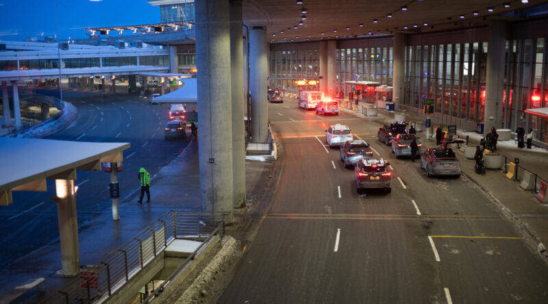 Airport terminal curbside with multiple cars and emergency flashing lights arriving or departing area near the entrance.