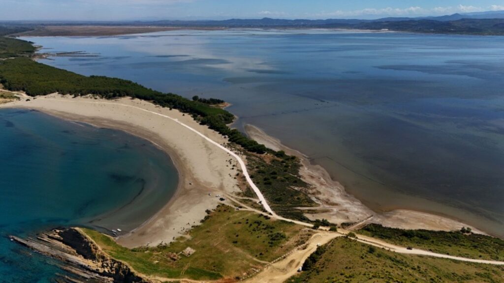 Aerial view of a crescent sandy beach along a narrow peninsula, with a white road winding through green scrub and trees; turquoise lagoon on the left and calm blue sea on the right.