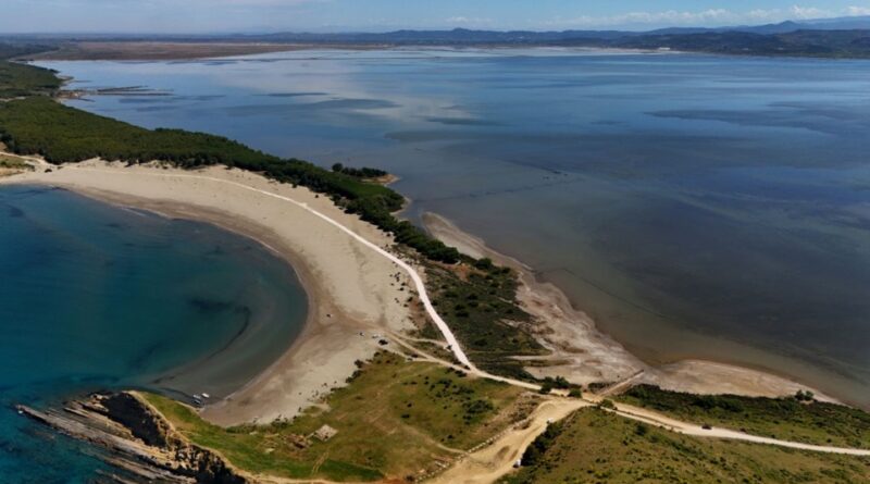 Aerial view of a crescent sandy beach along a narrow peninsula, with a white road winding through green scrub and trees; turquoise lagoon on the left and calm blue sea on the right.