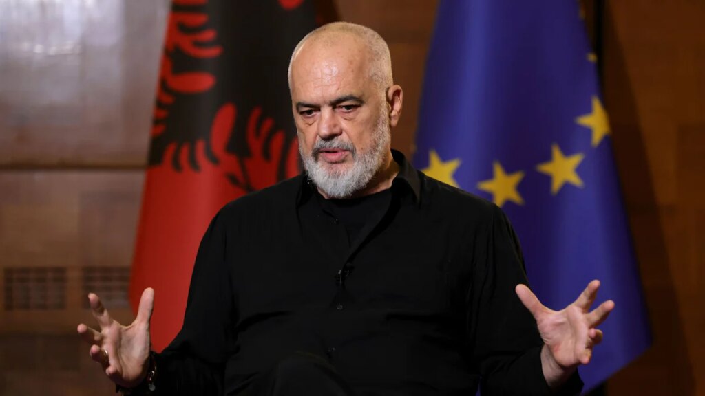Man with a gray beard in a black shirt speaking on a stage, with the Albanian flag and the EU flag in the background.
