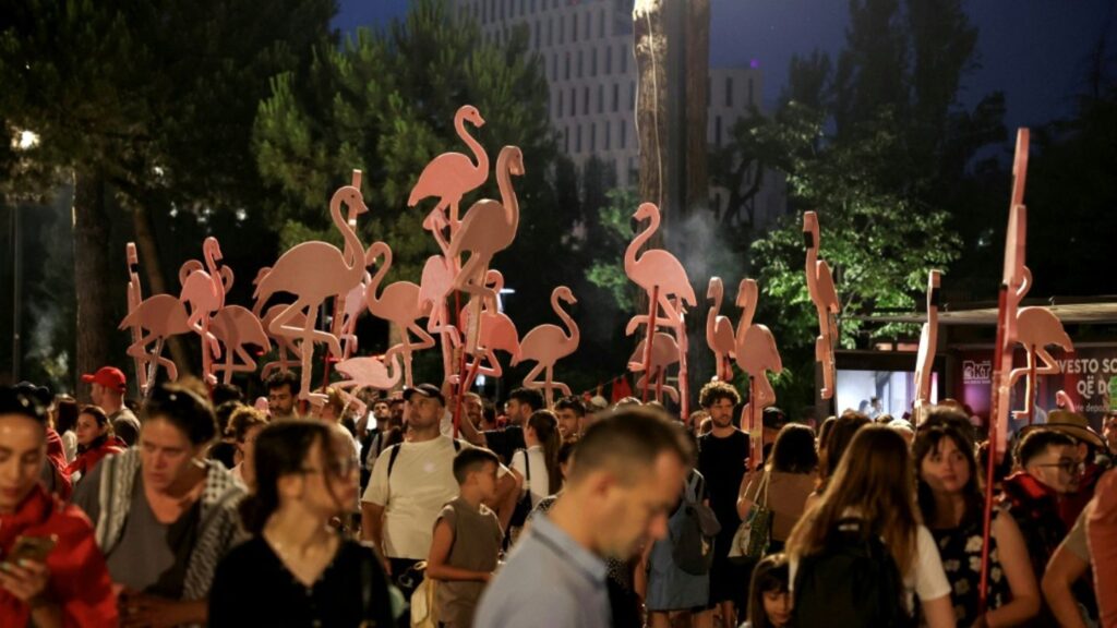 Nighttime festival crowd with tall pink flamingo cutouts on sticks above the attendees' heads by a park area.