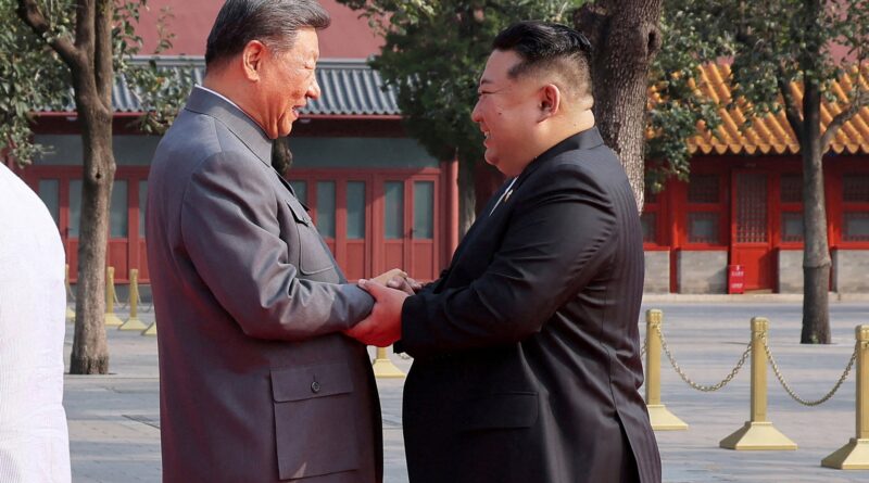 Two men in suits shake hands and smile at each other in a courtyard with red traditional buildings in the background.