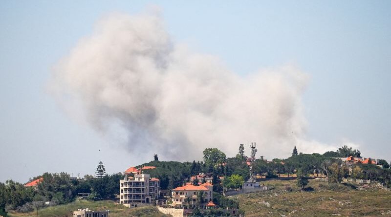 Smoke plume rising behind a hillside residential area with red-roofed houses and trees, obscuring the sky behind them.