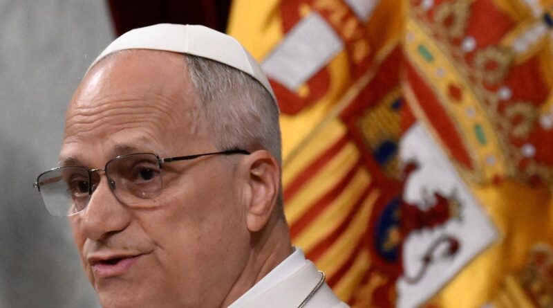 Side-profile of a clergyman wearing a white skullcap and glasses, with a colorful ceremonial banner in the background.