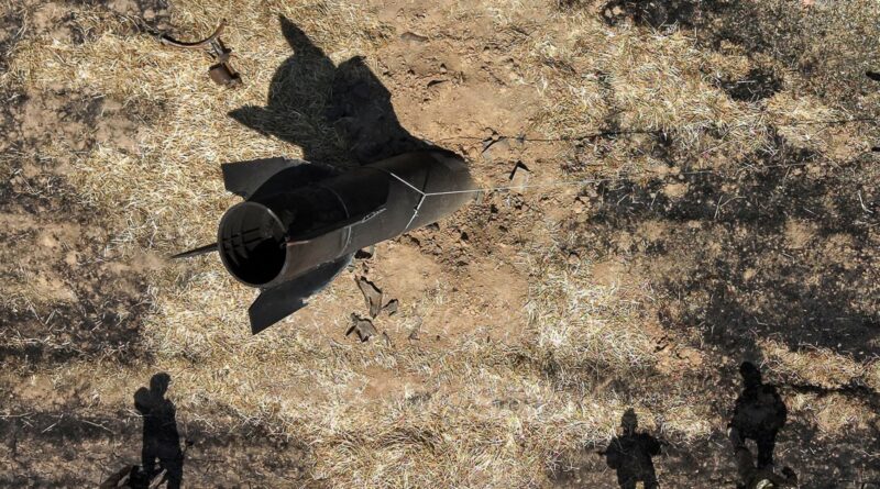 Overhead view of a black rocket resting on dry, dusty ground with shadowy silhouettes of people nearby.