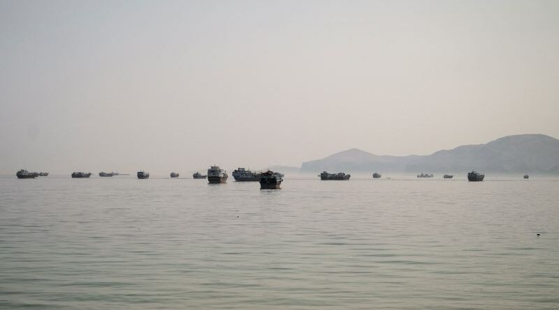 Calm sea with a line of small boats floating near the hazy horizon and distant hills.