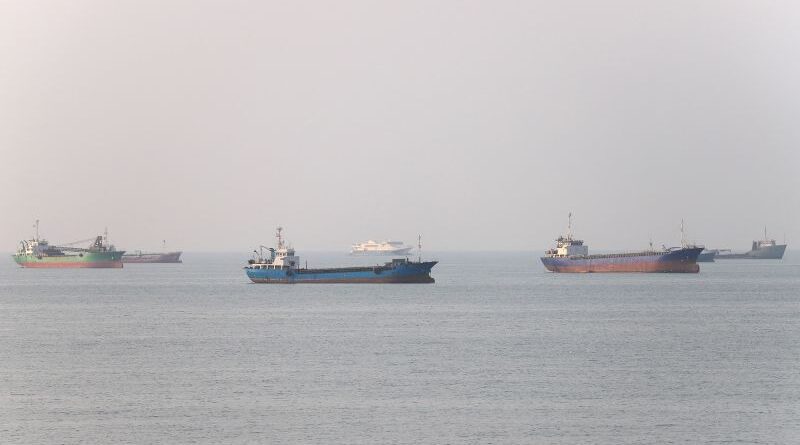 Several cargo ships floating on a calm sea under a hazy sky, with a distant horizon.