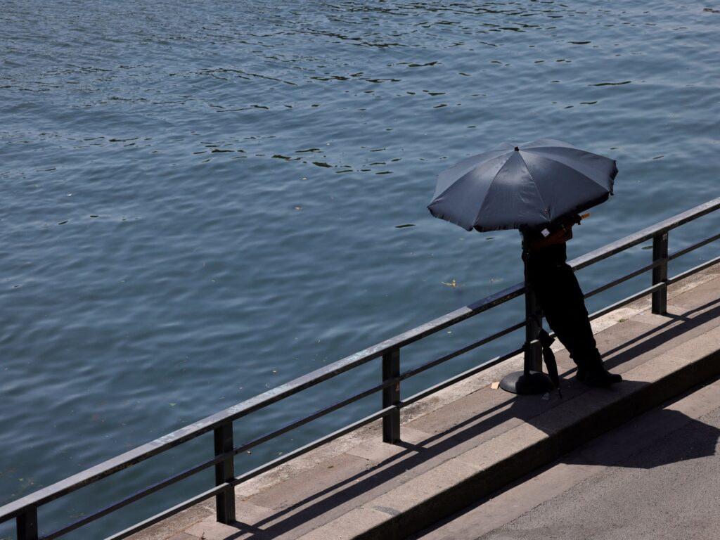 Person standing on a waterfront walkway holding a dark umbrella beside a metal railing over the water.