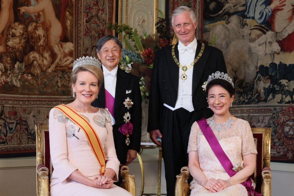 Two crowned women in elegant gowns with sashes sit on gilded chairs, flanked by two men in formal evening wear, against a richly patterned tapestry backdrop.