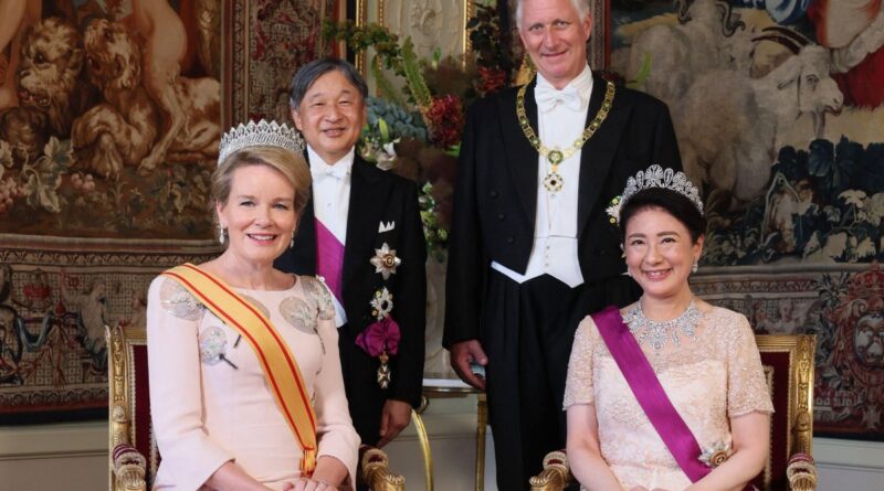 Two crowned women in elegant gowns with sashes sit on gilded chairs, flanked by two men in formal evening wear, against a richly patterned tapestry backdrop.