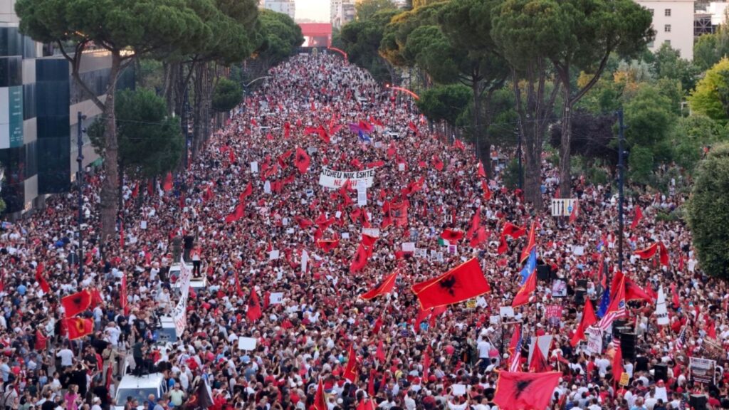 Massive street protest with thousands marching down a tree-lined avenue, many carrying red flags and signs.