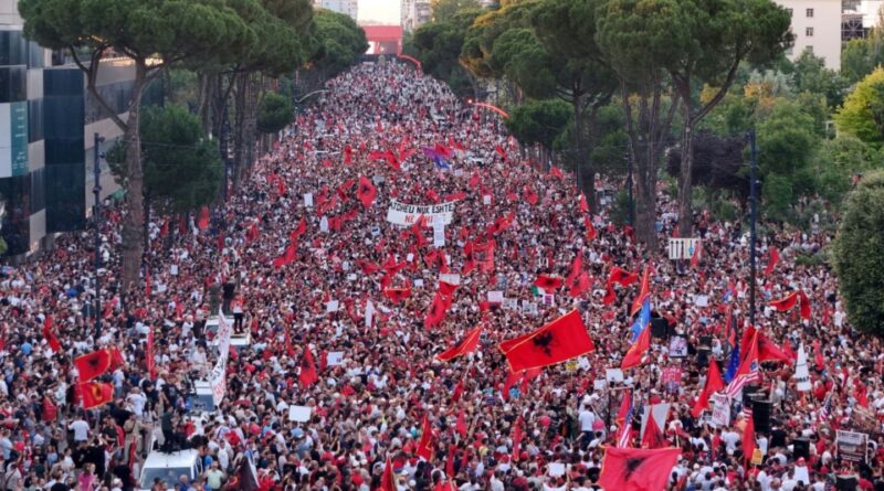 Massive street protest with thousands marching down a tree-lined avenue, many carrying red flags and signs.