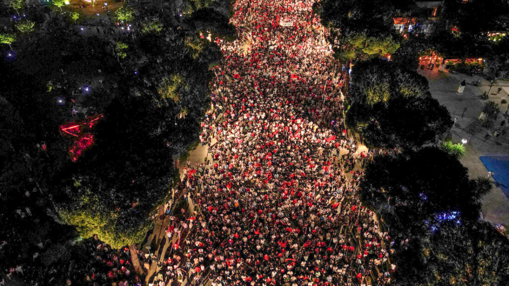 Aerial night view of a dense crowd marching down a city street, bordered by trees on both sides, with streetlights illuminating the scene.