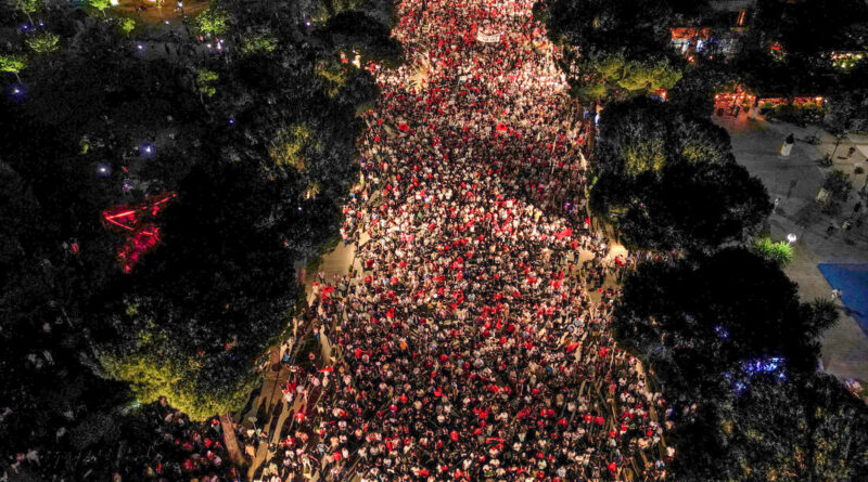 Aerial night view of a dense crowd marching down a city street, bordered by trees on both sides, with streetlights illuminating the scene.