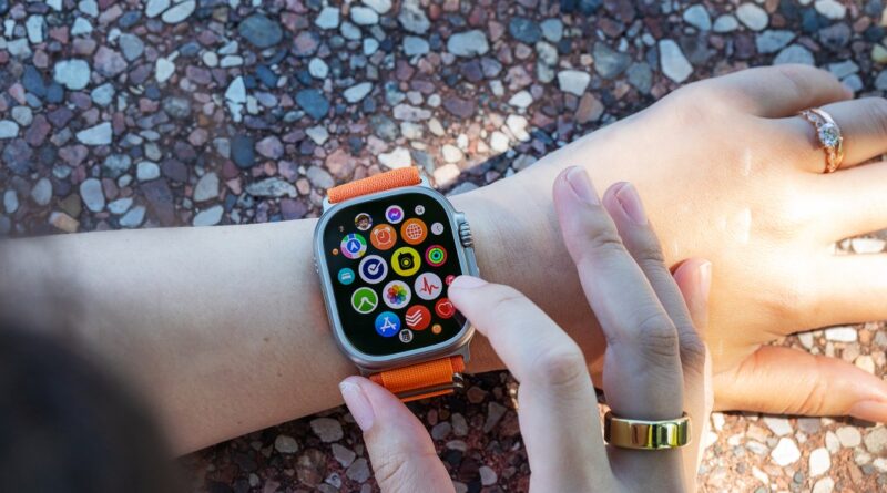 Person wearing an Apple Watch with an orange strap on their wrist, tapping the colorful app grid on the watch face outdoors over a rocky surface.