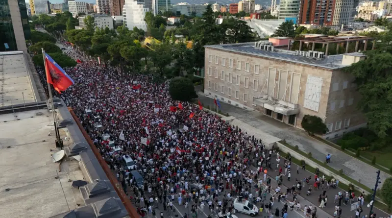 Large crowd of protesters gathered in a city plaza near a government building, waving red flags (Albania flag visible).