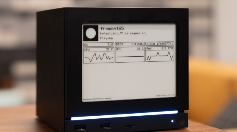 Small black mini PC with a monochrome screen showing system status and simple graphs on a desk lamp-lit table.
