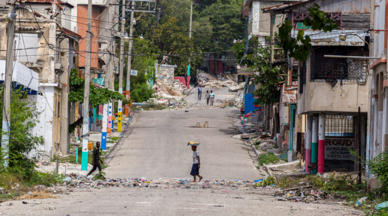 Ruined street in a disaster-hit town with rubble and damaged buildings; a person walks in the foreground with a basket on their head.
