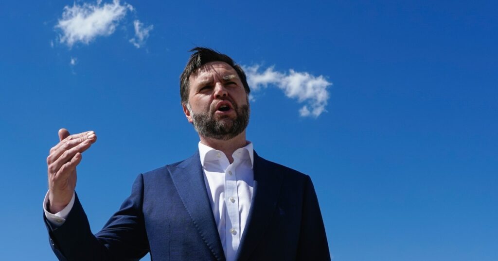 Man in a navy suit and white shirt delivering a speech outdoors against a clear blue sky, with one hand raised mid-gesture.