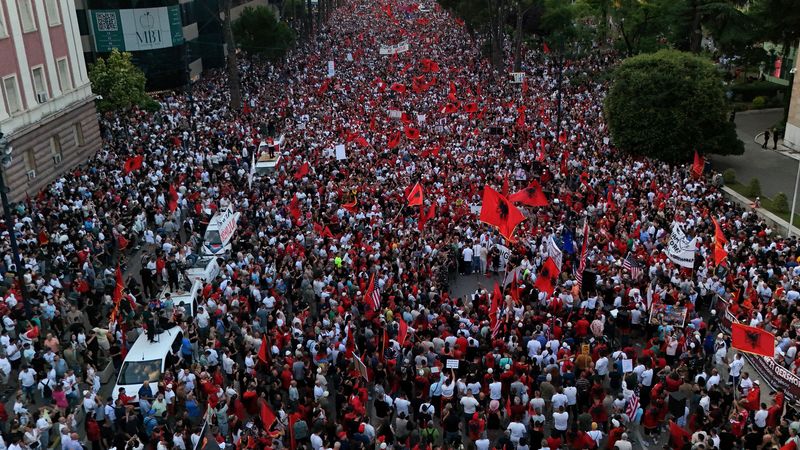 Aerial view of a dense street protest with thousands of people waving red flags and banners, crowd filling the avenue.