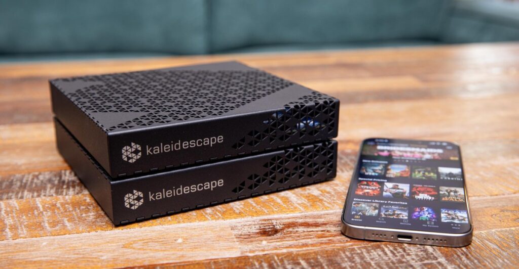 Two Kaleidescape media servers stacked on a wooden table beside a smartphone displaying a streaming app or movie library screen.