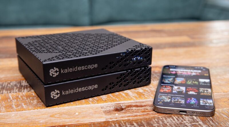 Two Kaleidescape media servers stacked on a wooden table beside a smartphone displaying a streaming app or movie library screen.