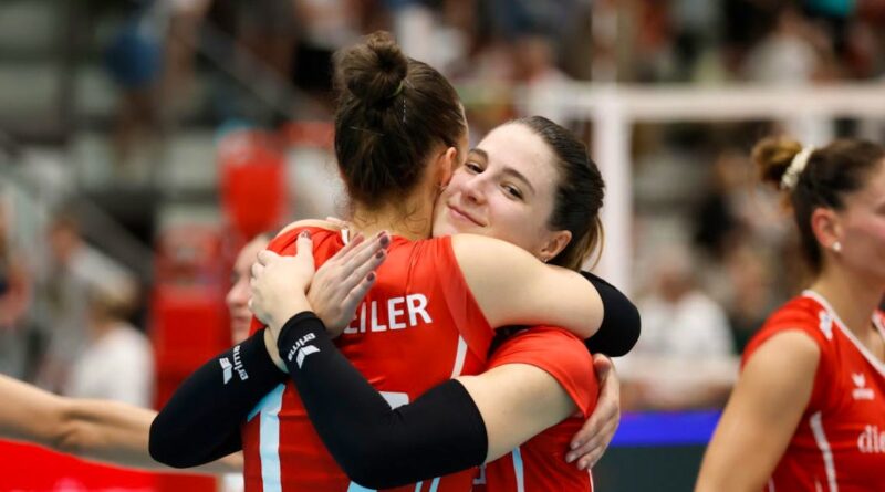 Two female volleyball players in red jerseys hug on the court, celebrating a point.